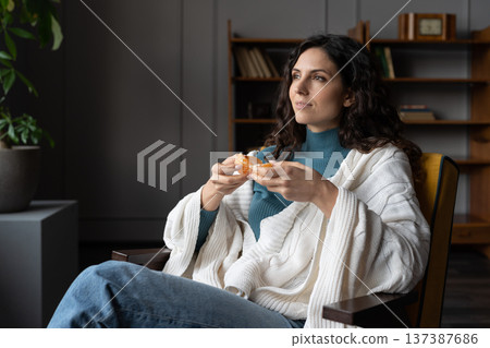 Young dreamy woman covered in knitted plaid peeling fresh tangerine orange while relaxing at home 137387686