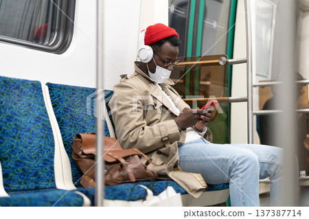 African hipster man in subway train wear face mask using cellphone listens to music with headphones. African hipster man in subway train wear face mask using cellphone listens to music with headphones. 137387714