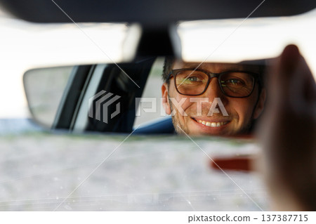 Cheerful man wearing glasses, adjusting mirror while sitting in car, looking in reflection Cheerful man wearing glasses, adjusting mirror while sitting in car, looking in reflection 137387715