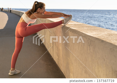 Fit sporty woman stretching legs, exercising near seaside road. Young female warm-up before workout Fit sporty woman stretching legs, exercising near seaside road. Young female warm-up before workout 137387719
