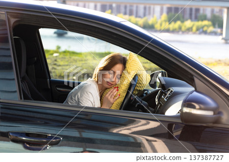 Tired woman driver asleep on pillow on steering wheel, resting after long hours driving car. Fatigue Tired woman driver asleep on pillow on steering wheel, resting after long hours driving car. Fatigue 137387727