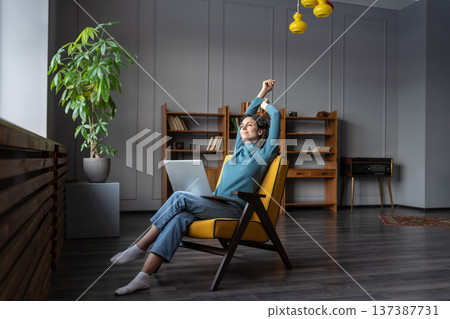Satisfied woman freelancer sitting on chair with laptop relaxing while working remotely from home Satisfied woman freelancer sitting on chair with laptop relaxing while working remotely from home 137387731