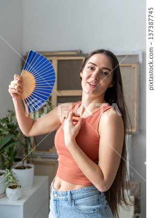 Positive smiling woman coping with overheating at home, posing at camera with hand paper fan. 137387745