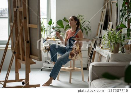 Concentrated woman painter painting picture on easel at creative home studio filled with plants. Concentrated woman painter painting picture on easel at creative home studio filled with plants. 137387746