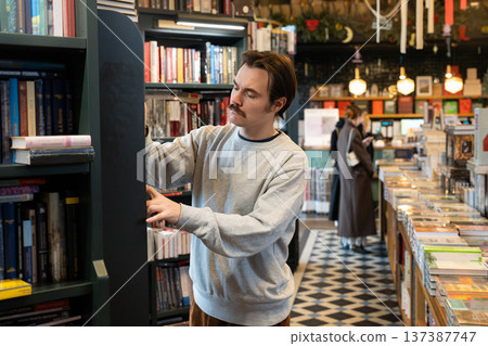 Intelligent focused man booklover searching for interesting books on shelf in cozy local bookstore. 137387747