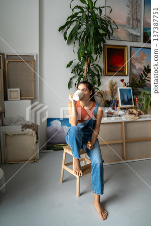 Painter woman relaxing with drink and sandwich while taking break at work on painting in studio. Painter woman relaxing with drink and sandwich while taking break at work on painting in studio. 137387751