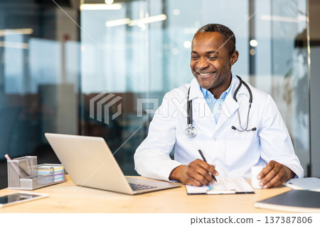 Doctor in lab coat and stethoscope smiling at desk, writing on a clipboard with laptop and tablet nearby, documenting patient care in a modern clinic workspace 137387806