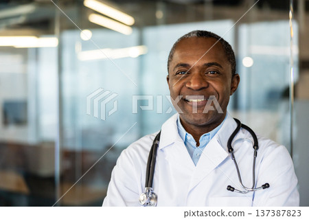 Mature african american male doctor in lab coat with stethoscope, smiling confidently for a professional portrait that conveys expertise, trust and compassionate patient care in clinic Mature african american male doctor in lab coat with stethoscope, smiling confidently for a professional portrait that conveys expertise, trust and compassionate patient care in clinic 137387823