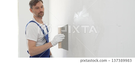 Male construction worker wearing protective gloves and blue construction coveralls, spreading plaster smoothly across wall using professional drywall taping knife during renovation project. Portrait Male construction worker wearing protective gloves and blue construction coveralls, spreading plaster smoothly across wall using professional drywall taping knife during renovation project. Portrait 137389983