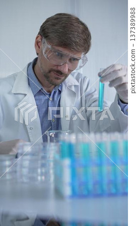 Male scientist researcher, wearing lab coat and protective glasses is looking to and examining test tub with blue liquid in modern laboratory, portrait view. Low key lighting Male scientist researcher, wearing lab coat and protective glasses is looking to and examining test tub with blue liquid in modern laboratory, portrait view. Low key lighting 137389988