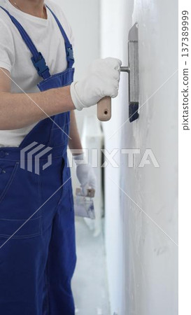 Professional construction worker wearing blue work overall is using a finishing trowel to carefully apply plaster on a wall, demonstrating expertise in home renovation Professional construction worker wearing blue work overall is using a finishing trowel to carefully apply plaster on a wall, demonstrating expertise in home renovation 137389999