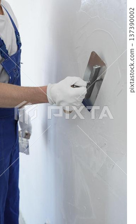 Professional construction worker wearing blue work overall is using a finishing trowel to carefully apply plaster on a wall, demonstrating expertise in home renovation 137390002