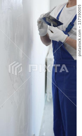 Professional construction worker wearing blue work overall is using a finishing trowel to carefully apply plaster on a wall, demonstrating expertise in home renovation Professional construction worker wearing blue work overall is using a finishing trowel to carefully apply plaster on a wall, demonstrating expertise in home renovation 137390005