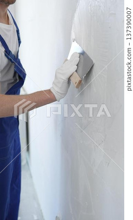 Man construction worker wearing protective gloves and blue construction coveralls, spreading plaster smoothly across wall using professional drywall taping knife Man construction worker wearing protective gloves and blue construction coveralls, spreading plaster smoothly across wall using professional drywall taping knife 137390007