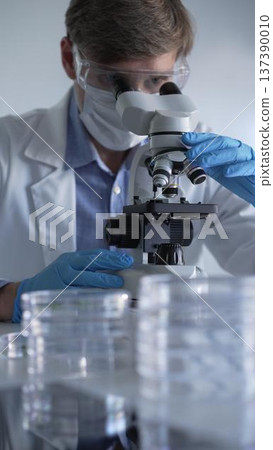 Man scientist researcher wearing blue gloves, protective glasses and medical mask is using microscope in modern laboratory, portrait view. Low key lighting photo. Science and Medicine concepts Man scientist researcher wearing blue gloves, protective glasses and medical mask is using microscope in modern laboratory, portrait view. Low key lighting photo. Science and Medicine concepts 137390010