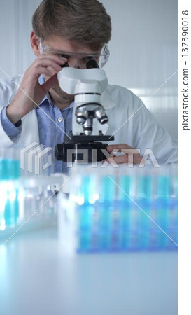 Male scientist researcher wearing lab coat and protective glasses, is using microscope in modern laboratory setup, portrait view. Low key lighting photo. Science concept Male scientist researcher wearing lab coat and protective glasses, is using microscope in modern laboratory setup, portrait view. Low key lighting photo. Science concept 137390018