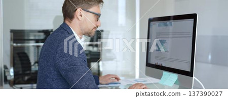 Businessman in a blue jacket analyzing financial data on computer, working in modern office, managing business strategy and reporting. Business people concept Businessman in a blue jacket analyzing financial data on computer, working in modern office, managing business strategy and reporting. Business people concept 137390027