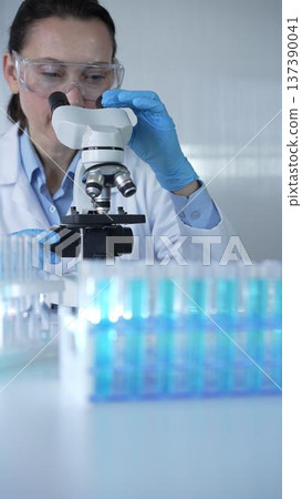 Female scientist wearing lab coat and protective blue gloves using microscope doing research in modern laboratory, portrait view. Low key lighting photo. Science and Medicine Female scientist wearing lab coat and protective blue gloves using microscope doing research in modern laboratory, portrait view. Low key lighting photo. Science and Medicine 137390041