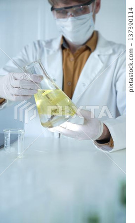 Male scientist wearing white protective gloves, mask and glasses is holding a yellow chemical solution inside an Erlenmeyer flask in a laboratory, vertical portrait. Science and medicine 137390114