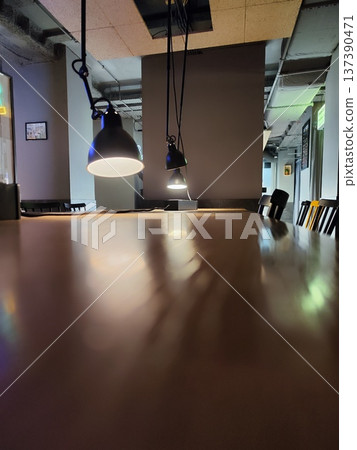 Long wooden table with reflections under three hanging black pendant lights in a dimly lit room. The ceiling has exposed ducts and a light-colored acoustic panel Long wooden table with reflections under three hanging black pendant lights in a dimly lit room. The ceiling has exposed ducts and a light-colored acoustic panel 137390471