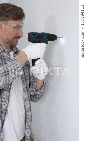 Middle aged man worker wearing grey checked shirt and white gloves, is drilling hole in a wall with cordless drill during home renovation work. Portrait view 137394113