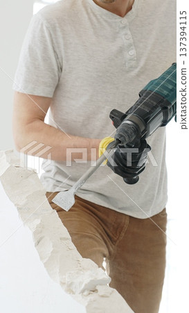 Unknown male construction worker with white t-shirt and protective gloves, is demolishing white wall with rotary hammer drill, generating debris on the floor, vertical close up view. Renovation Unknown male construction worker with white t-shirt and protective gloves, is demolishing white wall with rotary hammer drill, generating debris on the floor, vertical close up view. Renovation 137394115