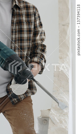 Close up of unknown male construction worker wearing beige checkered shirt and protective gloves, is demolishing white wall with rotary hammer drill, generating dust. Renovation project Close up of unknown male construction worker wearing beige checkered shirt and protective gloves, is demolishing white wall with rotary hammer drill, generating dust. Renovation project 137394118