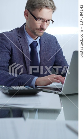 Businessman wearing blue suit is working with laptop, calculator and documents at the glass table in office set. Low lighting, vertical portrait. Business people and audit Businessman wearing blue suit is working with laptop, calculator and documents at the glass table in office set. Low lighting, vertical portrait. Business people and audit 137394142