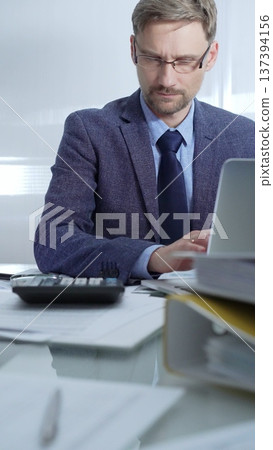Businessman wearing blue suit is working with laptop, calculator and documents at the glass table in office set. Low lighting, vertical portrait. Business people and audit Businessman wearing blue suit is working with laptop, calculator and documents at the glass table in office set. Low lighting, vertical portrait. Business people and audit 137394156