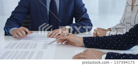 Business professionals analyzing contract details, gesturing with pens while discussing legal terms during collaborative meeting, focused on negotiation process 137394173