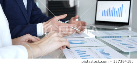 Business professionals around a glass table review reports and financial charts on laptops and printouts, gesturing and collaborating on strategy, analysis, and planning Business professionals around a glass table review reports and financial charts on laptops and printouts, gesturing and collaborating on strategy, analysis, and planning 137394193