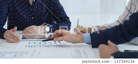 Professional colleagues examining financial documents, studying performance charts on paper and digital tablet during collaborative meeting in modern workplace setting 137394216