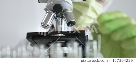 Scientist wearing green gloves and a face mask selecting a test tube with transparent liquid from a rack near a microscope in a lab. Medicine, healthcare and science concept 137394239
