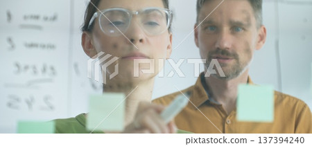 Professional woman with glasses marking glass board, arranging sticky notes while male colleague reviews strategic planning within modern, well lit workspace. Business people concept 137394240