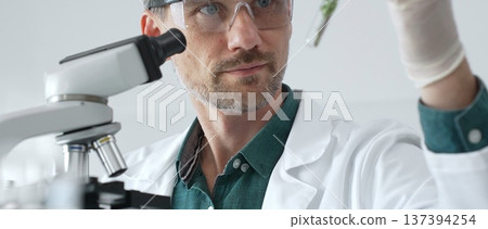Adult male scientist in a lab coat and gloves examining a plant sample in a test tube, with a microscope set up in the foreground. Medicine, healthcare and science concept Adult male scientist in a lab coat and gloves examining a plant sample in a test tube, with a microscope set up in the foreground. Medicine, healthcare and science concept 137394254