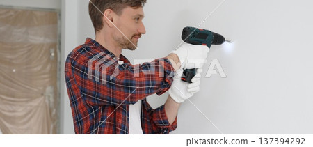 Middle aged male worker wearing red checked shirt and white gloves, is drilling hole in a wall with cordless drill during home renovation work. Portrait view 137394292