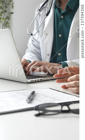 Male doctor wearing lab coat over a green shirt, is typing on a laptop and listening to a woman patient during medical consultation in clinic. Medicine and health care Male doctor wearing lab coat over a green shirt, is typing on a laptop and listening to a woman patient during medical consultation in clinic. Medicine and health care 137394308
