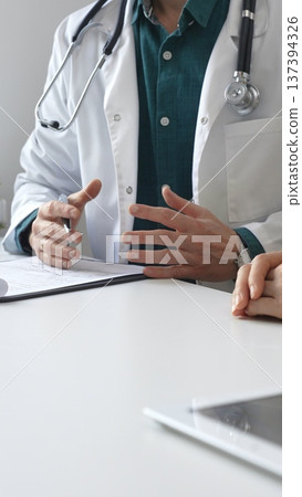Doctor man wearing medical coat over green shirt, and stethoscope is gesturing with hands over white table and medical records while explaining diagnosis to patient. Medicine and health care concept Doctor man wearing medical coat over green shirt, and stethoscope is gesturing with hands over white table and medical records while explaining diagnosis to patient. Medicine and health care concept 137394326