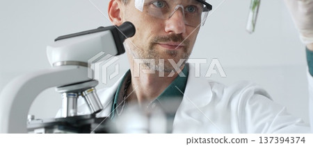 Adult male scientist in a lab coat and gloves examining a plant sample in a test tube, with a microscope set up in the foreground. Medicine, healthcare and science concept Adult male scientist in a lab coat and gloves examining a plant sample in a test tube, with a microscope set up in the foreground. Medicine, healthcare and science concept 137394374