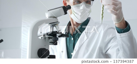 Masked adult man researcher examining green botanical samples in test tube in a sterile laboratory during biological analysis. Medicine, healthcare and science concept Masked adult man researcher examining green botanical samples in test tube in a sterile laboratory during biological analysis. Medicine, healthcare and science concept 137394387