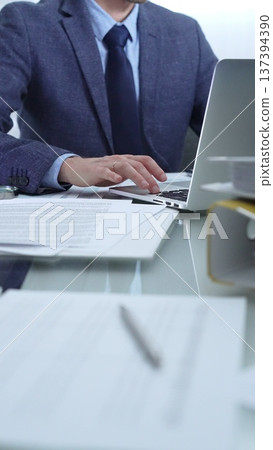 Businessman wearing blue suit is working with laptop, calculator and documents at the glass table in office set. Low lighting, close-up vertical view. Business people and audit 137394390