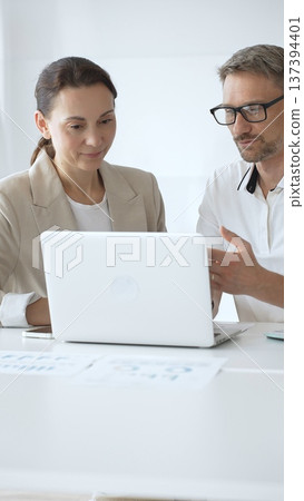 Businessman and adult businesswoman reviewing marketing analytics on laptops, working together strategically in contemporary workplace with minimalist interior design and collaborative atmosphere 137394401