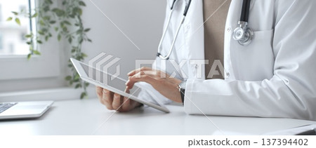 Doctor man wearing lab coat and stethoscope using digital tablet in modern medical office, browsing patient information or conducting online consultation. Medicine and health care 137394402