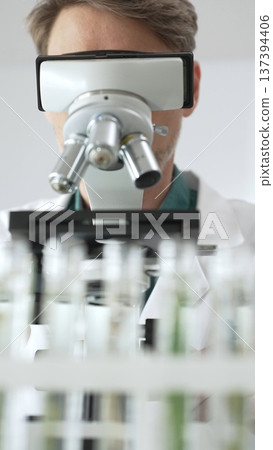 Man scientific researcher in protective gear analyzing specimens through a microscope in a sterile laboratory environment. Medicine, health care and science concept Man scientific researcher in protective gear analyzing specimens through a microscope in a sterile laboratory environment. Medicine, health care and science concept 137394406