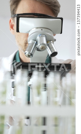 Man scientific researcher in protective gear analyzing specimens through a microscope in a sterile laboratory environment. Medicine, health care and science concept Man scientific researcher in protective gear analyzing specimens through a microscope in a sterile laboratory environment. Medicine, health care and science concept 137394413