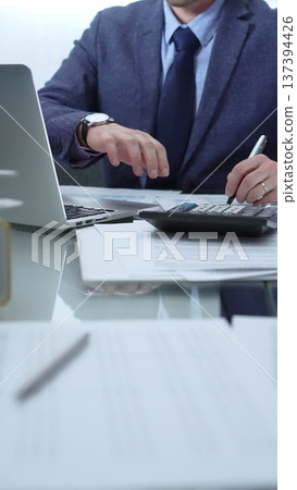 Businessman wearing blue suit is working with laptop, calculator and documents at the glass table in office set. Low lighting, close-up vertical view. Business people and audit Businessman wearing blue suit is working with laptop, calculator and documents at the glass table in office set. Low lighting, close-up vertical view. Business people and audit 137394426
