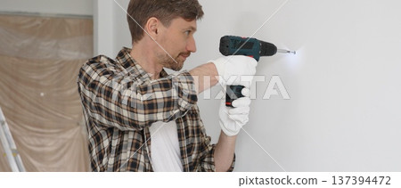 Middle aged man wearing checked shirt and white gloves, is drilling hole in a wall with cordless drill during home renovation work. Portrait view Middle aged man wearing checked shirt and white gloves, is drilling hole in a wall with cordless drill during home renovation work. Portrait view 137394472