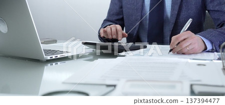 Businessman or accountant wearing blue suit is working with laptop, calculator and documents in office. Low lighting, close-up view. Business people, audit concept 137394477