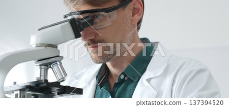 Adult male scientist wearing protective glasses and lab coat analyzing samples with a microscope in a modern laboratory setting. Medicine, healthcare and science concept Adult male scientist wearing protective glasses and lab coat analyzing samples with a microscope in a modern laboratory setting. Medicine, healthcare and science concept 137394520