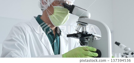 Medical scientist wearing protective mask, cap and green gloves using microscope doing scientific research in laboratory, developing vaccine or medication. Medicine, healthcare and science concept Medical scientist wearing protective mask, cap and green gloves using microscope doing scientific research in laboratory, developing vaccine or medication. Medicine, healthcare and science concept 137394536